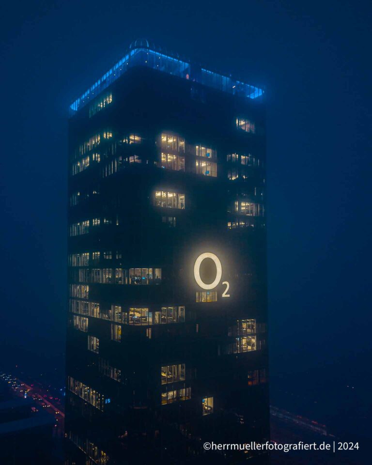 Die Spitze des o2 Towers München mit blau beleuchteter Sky Lounge und o2 Logo